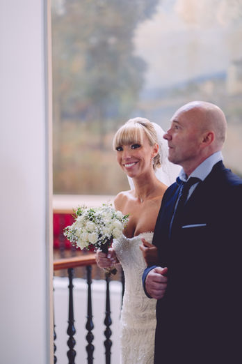 Mariée souriante avec bouquet blanc, accompagnée d’un homme élégant lors de son entrée dans la salle de cérémonie de mariage.