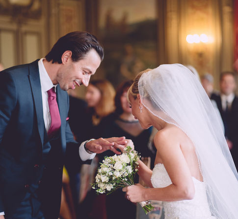 Échange des alliances entre la mariée et le marié dans une salle de cérémonie élégante, bouquet de fleurs blanches à la main.