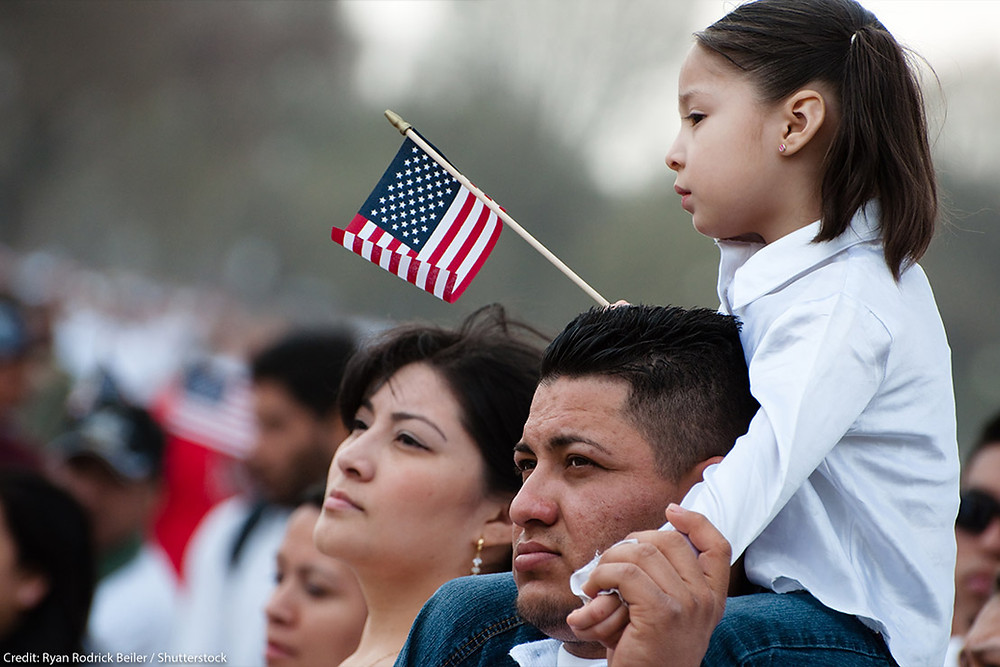 A young child sitting on a man's shoulders at an immigration event, waving a small United States flag.