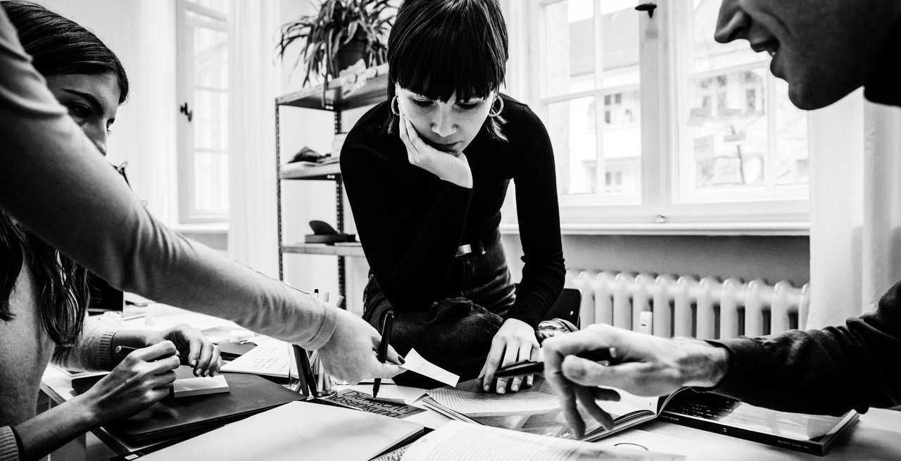 Black-and-white team meeting around a table, one woman leans over papers while others point at notes in a bright office.