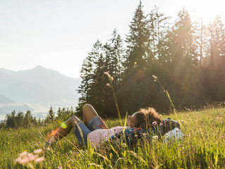 A Black woman lies in a field as the sun sets behind trees, with mountains in the distance.