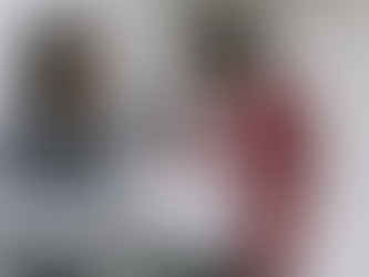 Two people at a glass table, engaged in discussion. One in a plaid shirt listens, the other in a red blazer points at documents. Bright room.