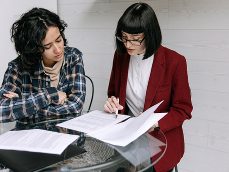 Two people at a glass table, engaged in discussion. One in a plaid shirt listens, the other in a red blazer points at documents. Bright room.