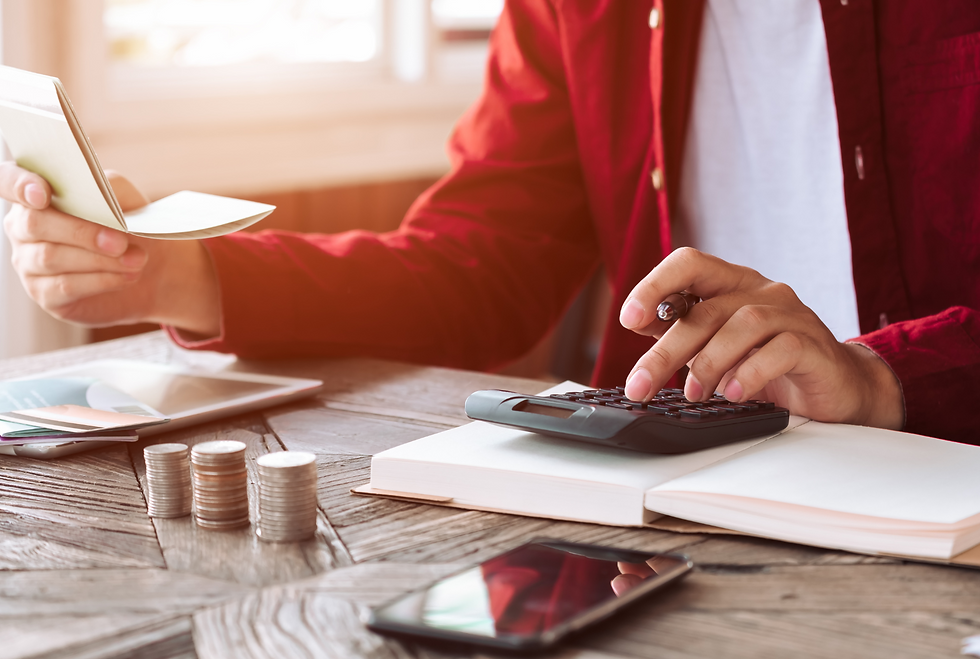 Man in red shirt using a calculator while holding a checkbook, with stacks of coins, a smartphone, and a tablet on a wooden table.