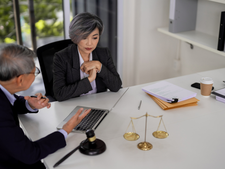 Two professionals in a meeting at an office desk with a laptop, scales, gavel, and documents. One gestures while the other listens pensively.
