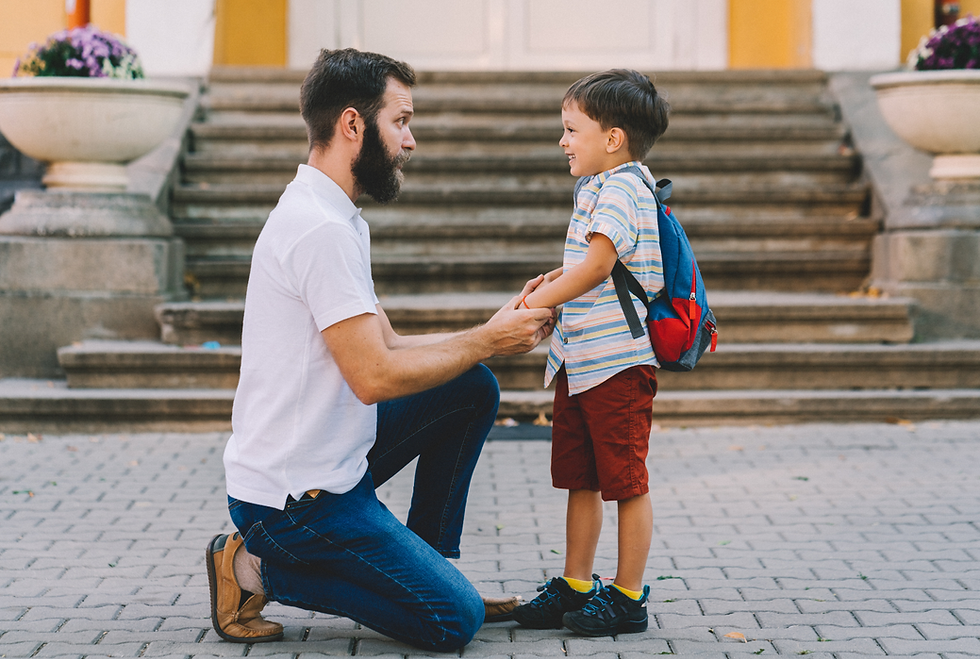 Man kneels, holding hands with smiling boy in striped shirt and red shorts, wearing a blue backpack. They're in front of stone steps.