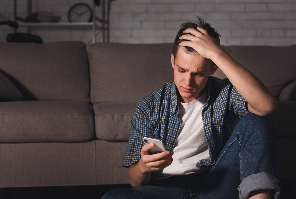 Man in casual clothes sits on the floor, holding a phone, looking worried. Gray sofa in the background, dimly lit room creating a tense mood.