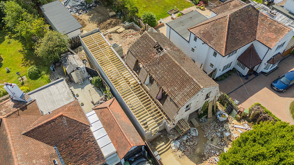 Aerial view of a house renovation with exposed roof beams, rubble, ladders, and tiled homes around a sunny suburban yard.