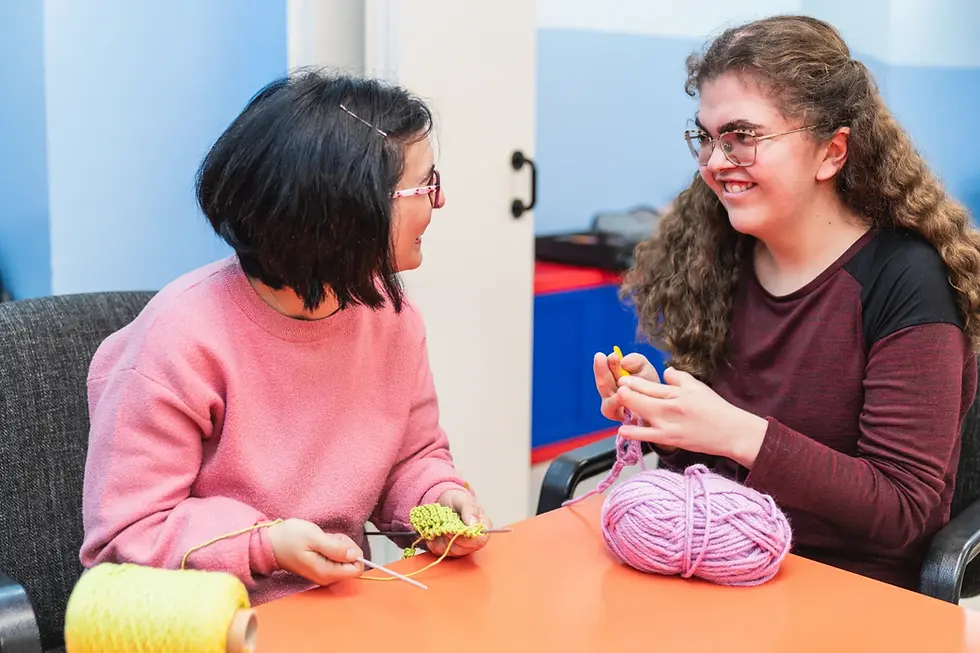 Two young women with disability sitting next to each other smiling whilst practising their knitting.