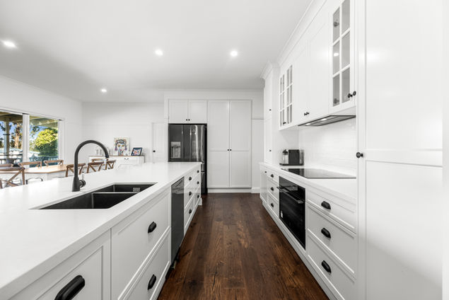 Long view of white kitchen with black taps and timber flooring