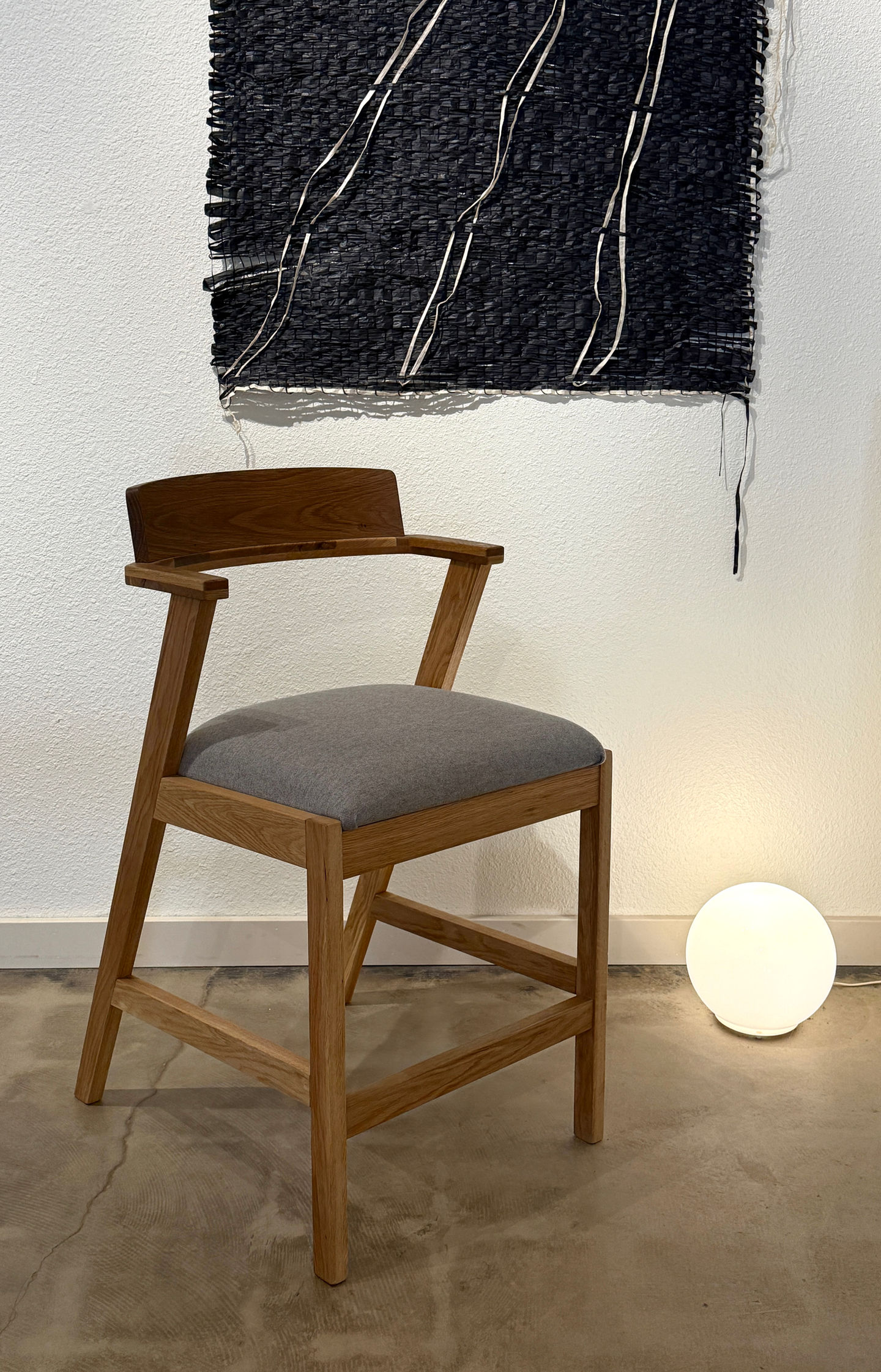 A modern oiled Walnut dining chair with unique half arms and a zigzag shape with soft grey flannel upholstered seat, on a concrete floor against a white wall