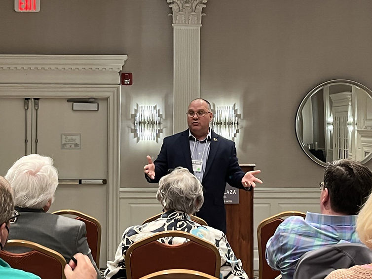 Steve Holden in a dark suit speaks at a podium in front of a group of constituents.