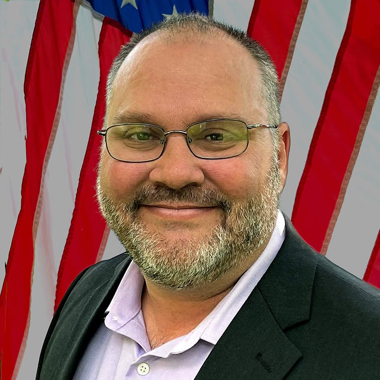 Steve Holden smiling in front of an American flag, wearing glasses, a light purple dress shirt, and a dark blazer.
