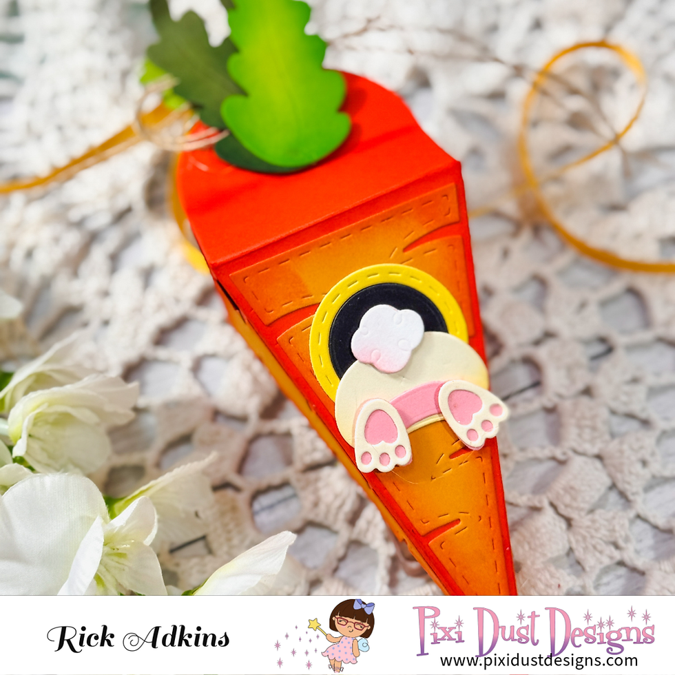 Cartoon carrot-shaped box with bunny feet and tail on lace fabric. White flowers nearby. Text: Rick Adkins, Pixi Dust Designs.