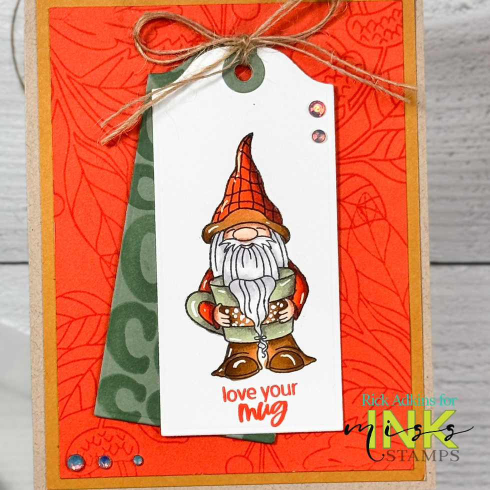 Gnome holding a mug on a gift tag, with "love your mug" text. Red and green leaf-patterned background, twine bow, and colorful gems.