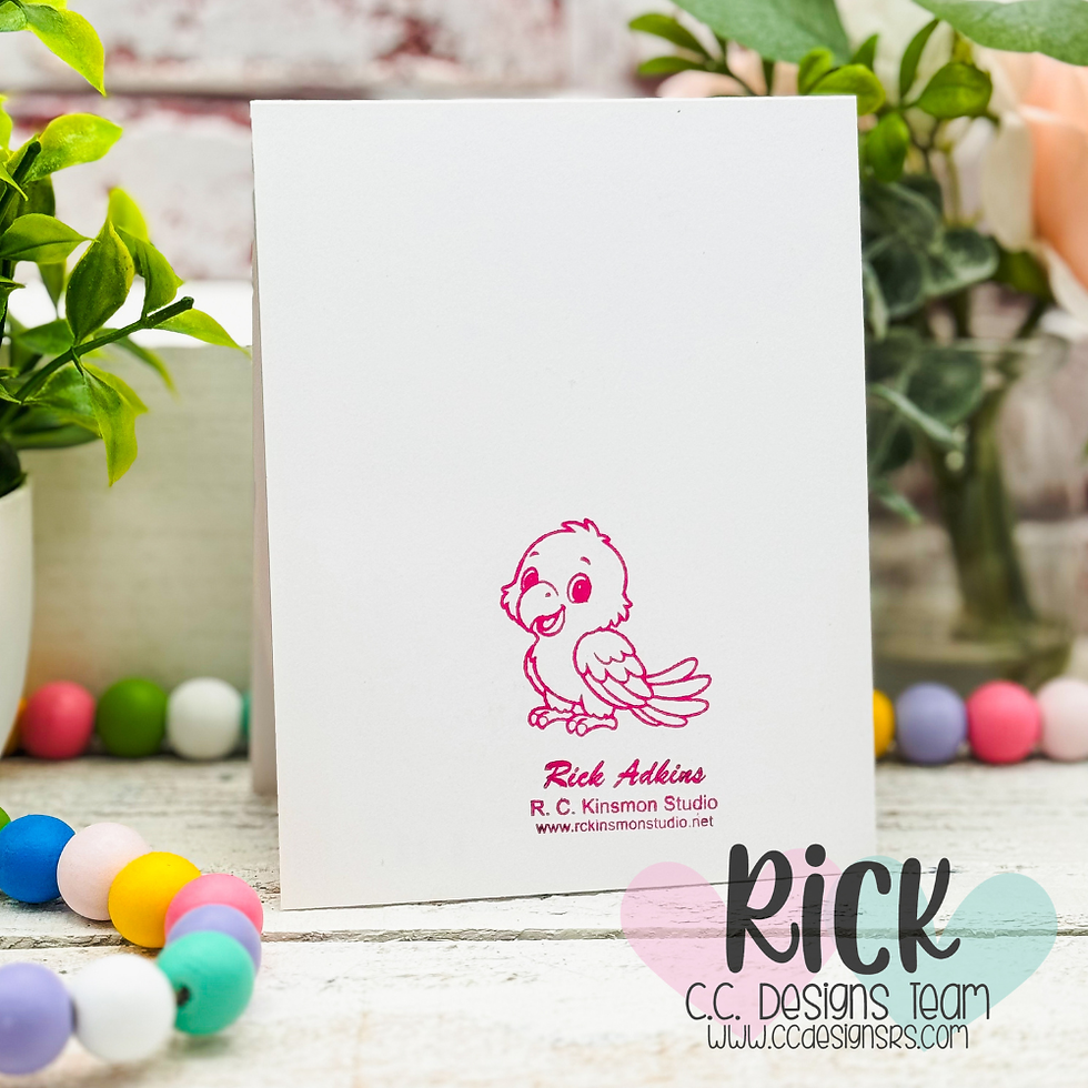 Blank white greeting card with pink bird logo and Rick Adkins text, styled among green plants and pastel beads on a rustic table