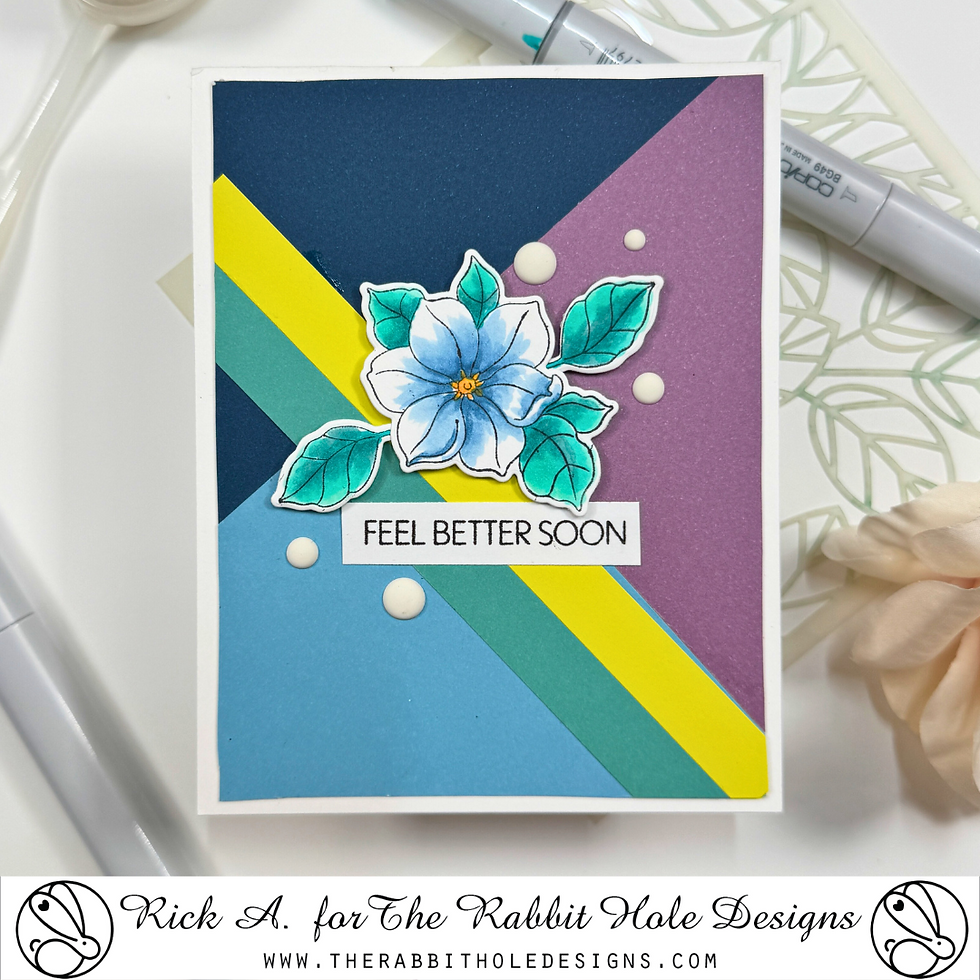 Greeting card with blue flower on colorful geometric background. Text: "FEEL BETTER SOON." Pens and stencil visible in the background.