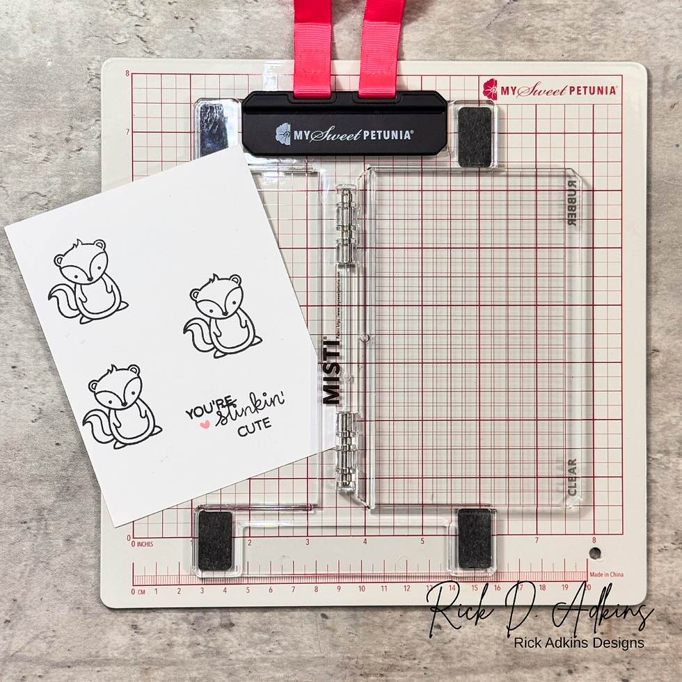 Cardmaking tool with grid lines and pink accents. A card with three cartoon skunks and the text "You're stinkin' cute" is placed on it.