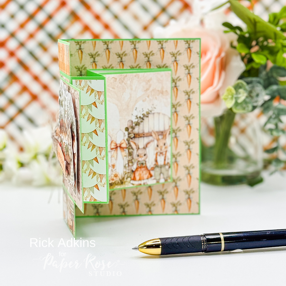 Handmade card with rabbits in orange attire against a carrot-patterned background. Pen nearby. Text: "Rick Adkins for Paper Rose Studio."