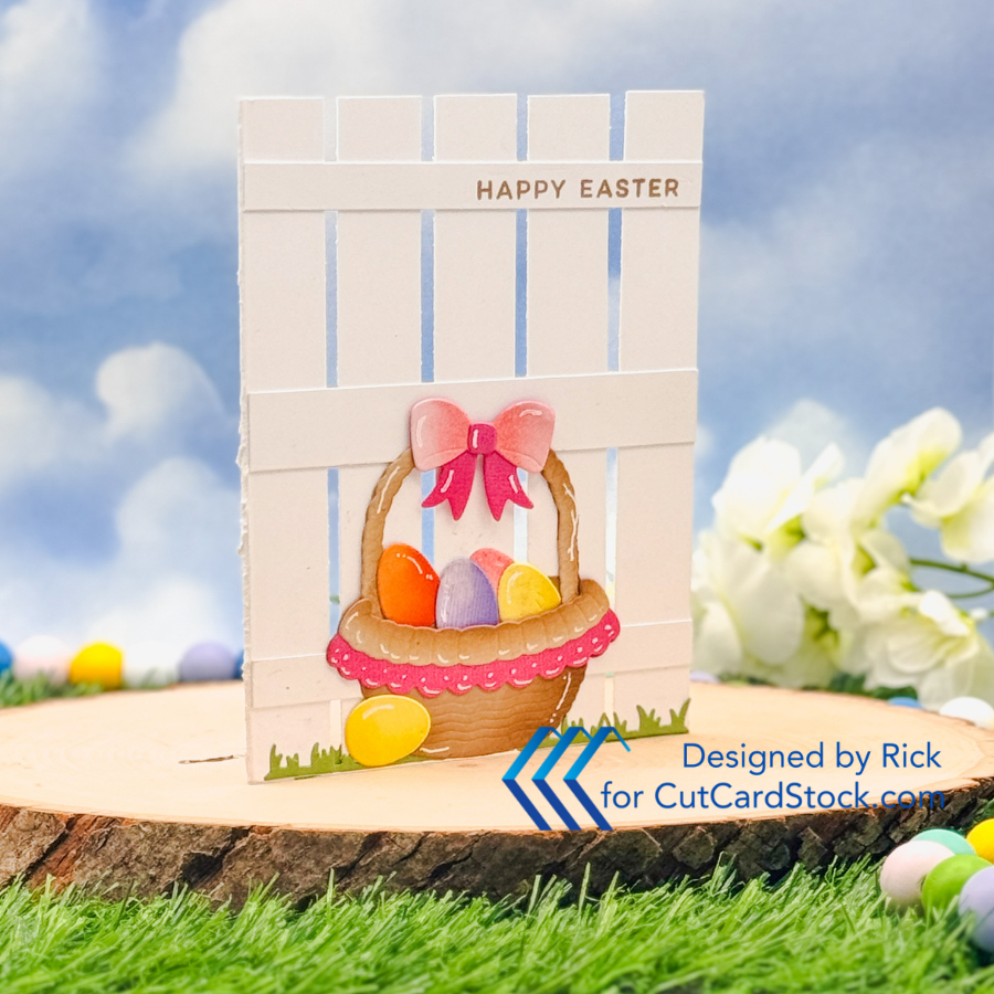 Easter card with a white fence, basket of colorful eggs, and pink bow. Text reads Happy Easter. Background has grass and flowers.