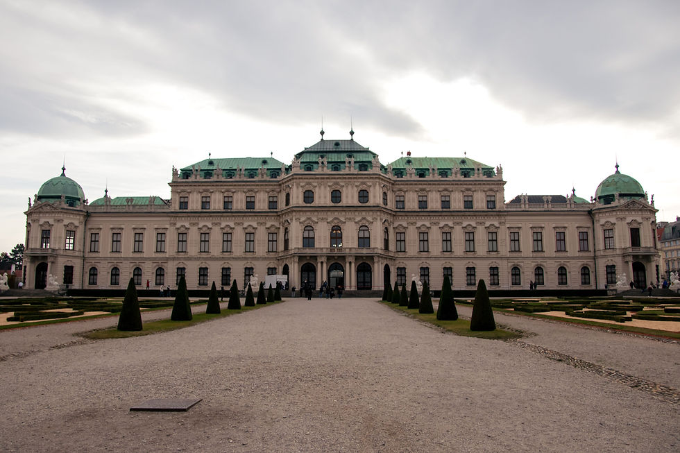 Grand historic palace with green domes and formal gardens, viewed from a gravel path under a cloudy sky.