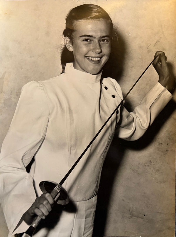 A smiling young woman in a white fencing uniform holds a foil. The background is a plain wall, creating a vintage black-and-white atmosphere.
