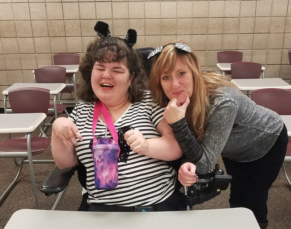 Me and my assistant smiling in a classroom, me in a wheelchair, wearing striped shirts and animal ear headbands. This was our first Halloween in college and I was unsure if people would dress up, so we played it safe with zebra ear headbands.