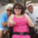 Three smiling people pose outdoors, a young woman in a pink dress in a wheelchair flanked by two older adults in sunglasses. I am sandwiched between my grandparents . this picture exudes their love for me.