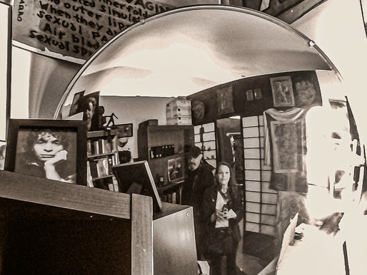 Reflection in a round mirror of a man and woman in a room with bookshelves. Sepia tone, curly-haired portrait, and text on posters visible.
