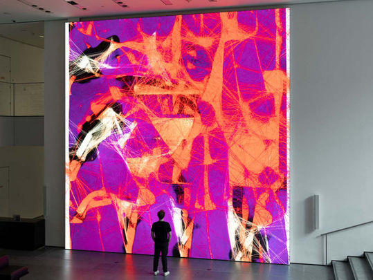 A person stands in front of a large abstract digital art piece with vivid purple and orange patterns, in a modern gallery setting.