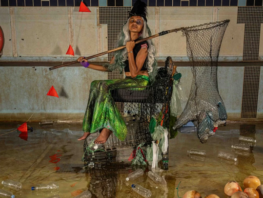 A person in a mermaid costume fishes with a net in a water-filled room. A float reads "Water for Sale." Plastic bottles float around.