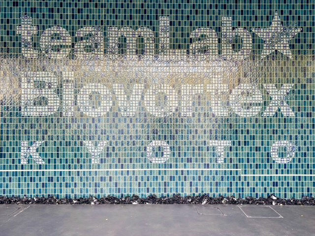 Mosaic wall with "teamLab★ Biovortex KYOTO" text in white against a green and blue tiled background, creating a modern and artistic feel.
