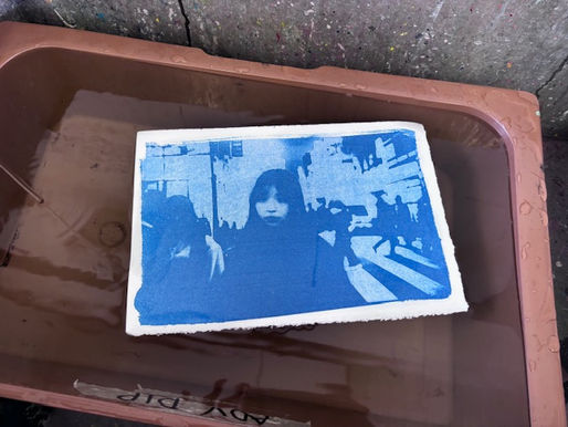 A blue-toned photo in a developing tray shows people walking in a city scene. The tray is brown and sits on a concrete surface.