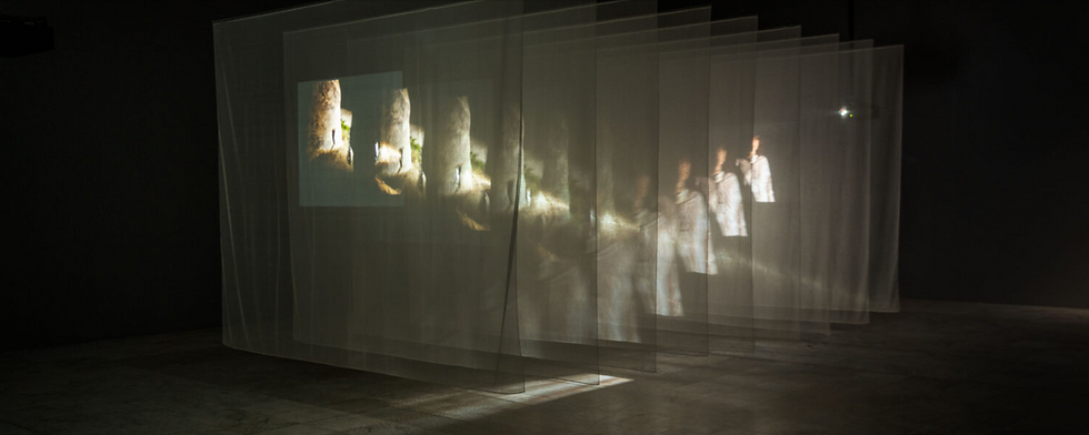 Projected images on layered translucent screens in a dim room create a ghostly effect. Figures and landscapes blend with ambient light.
