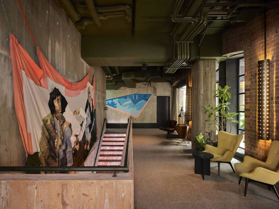 Dimly lit hallway with modern art on concrete walls, including a large tapestry and a blue geometric mural. Yellow chairs and potted plants add color.