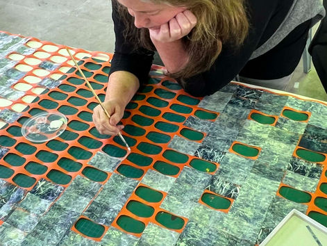 Woman leans over a table, carefully painting an orange lattice over green collage pieces with a thin brush in a craft room.