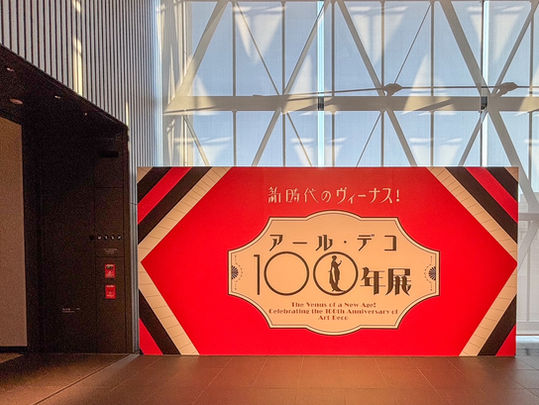 Red Art Deco exhibition sign with geometric patterns, celebrating 100th anniversary. Glass-paneled background in a brightly lit space.