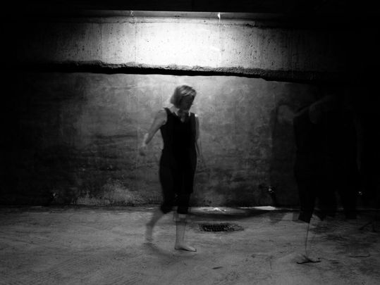 A blurred figure in black dances in an empty, dimly lit concrete room, creating a ghostly and mysterious atmosphere.