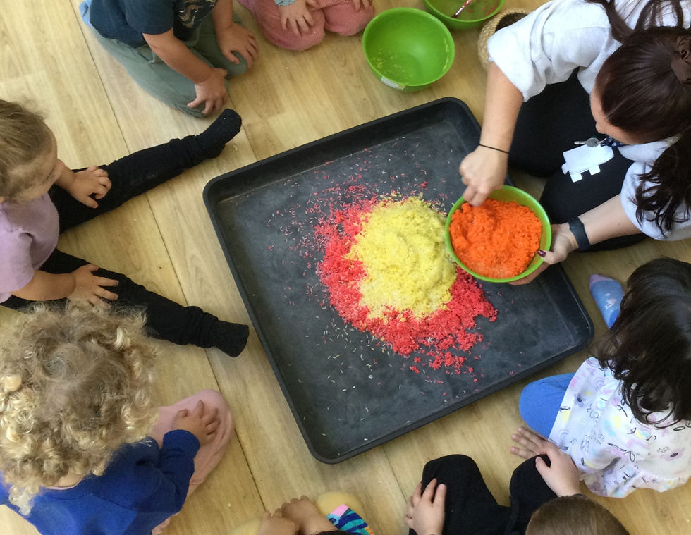 Children and an adult sit on a wooden floor around vibrant colored rice in a tray. Bright red, orange, and yellow hues fill the scene.