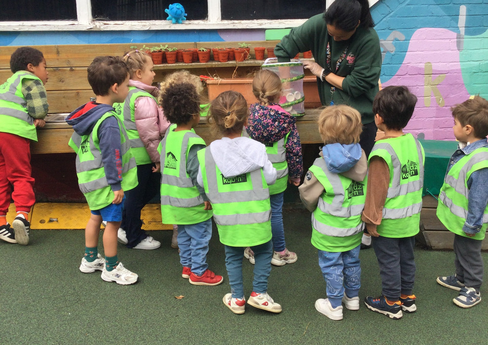 A group of children in the green room at Kidz Kabin Pembroke Studios nursery in Muswell Hill are outside with their teacher and learning about the lifecycle of a caterpillar