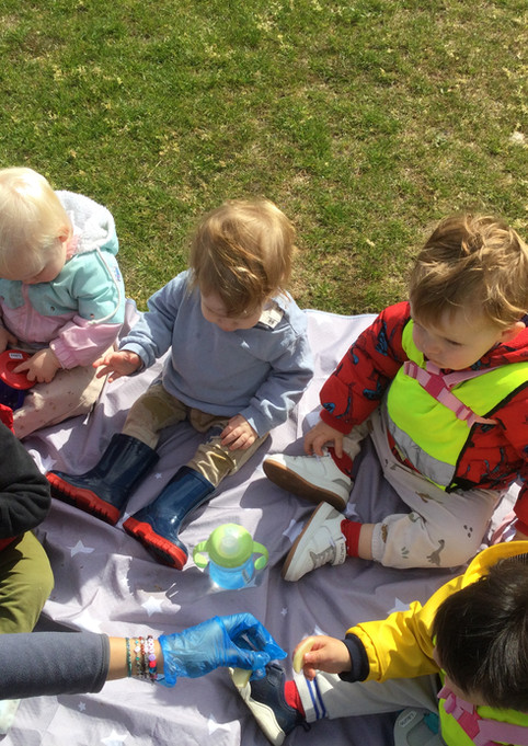 A group of blue room children from Kidz Kabin Shropshire Hall in Wood Green are sitting on a picnic blanket in the park enjoying snack time
