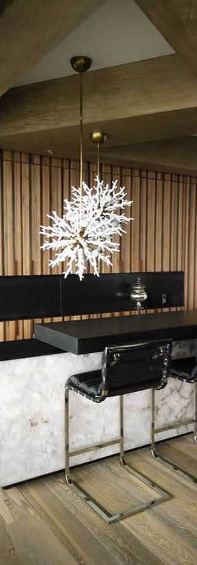 Barra de bar con acabado de mármol blanco junto con superficies negras brillantes. Hay 3 sillas altas de metal tapizadas con un material de apariencia de piel color negro. Sobre la barra hay 2 lámparas decorativas en forma de copos de nieve. Toda la pared del fondo cuenta con páneles de madera.