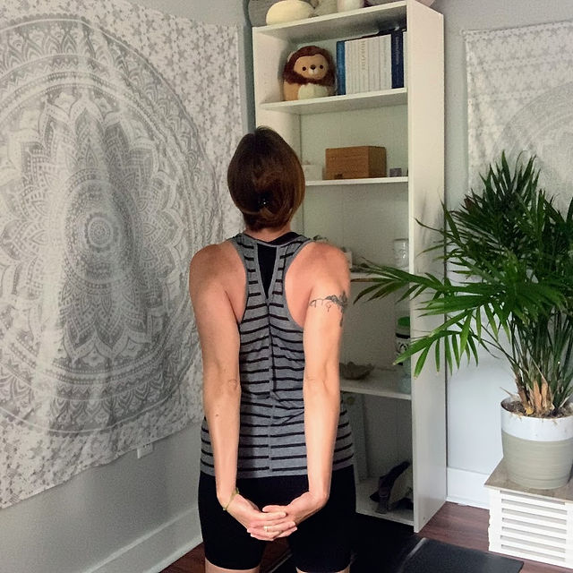 back view of a women in grey and black striped yoga shirt demonstrating 4th pose, hands clasped behind back