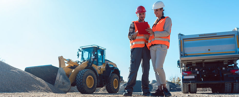 Two construction workers in hard hats and orange vests review a clipboard. A bulldozer and dump truck are in the sunny, gravel-filled site.