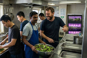 Ship cook preparing fresh vegetables in a commercial vessel kitchen with onboard growing system in the background, highlighting fresh food production at sea