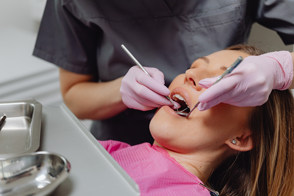 Dentist wearing gloves examines a woman's teeth with tools. The woman wears a pink dental bib, lying back in a clinical setting.