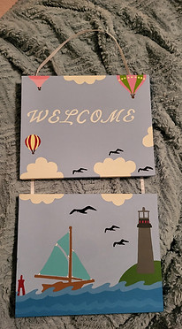Two canvases attached by ribbon, that are painted with balloons, clouds, birds, a sailboat and a lighthouse, with the word Welcome on one of the canvases.