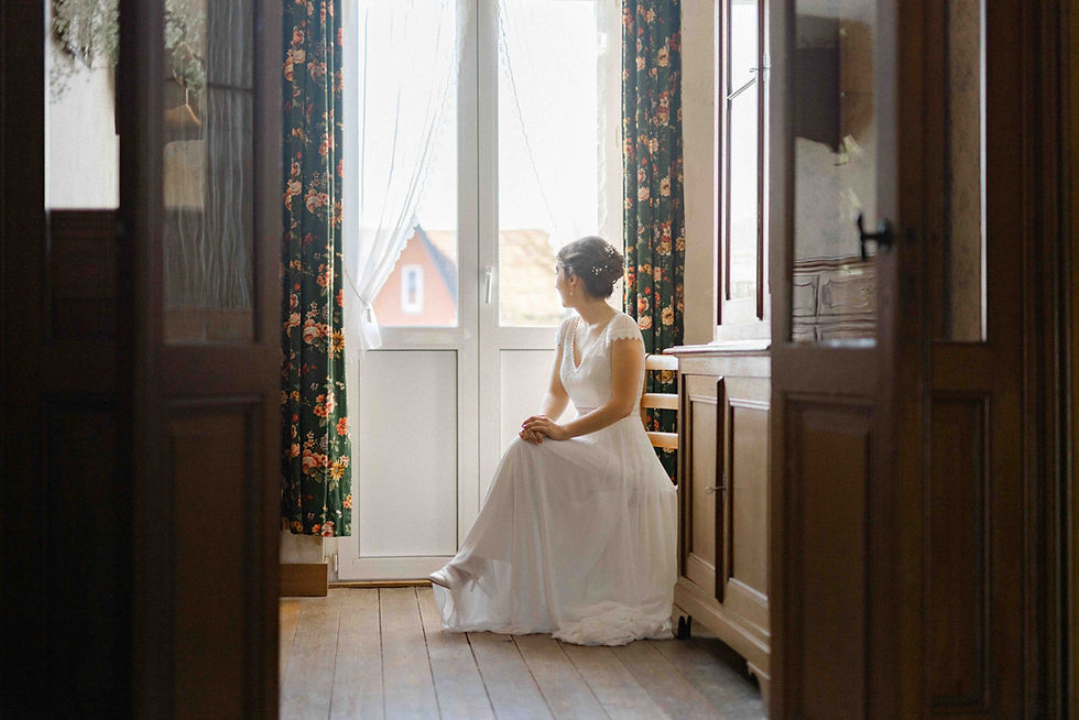 Mariée assise près d'une fenêtre baignée de lumière douce, regard tourné vers l’extérieur, illustrant