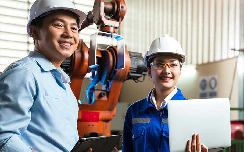 Two smiling precision engineering professionals in hard hats observing an automated robotic arm