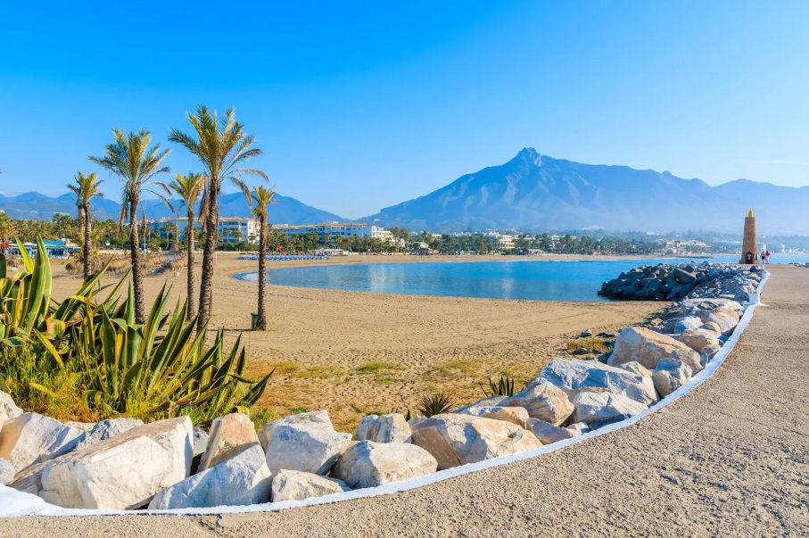 Marbella tengerpart, stány szélén nagy fehér kövek, háttérben öt pálmafa, a kék tenger és a hegyek